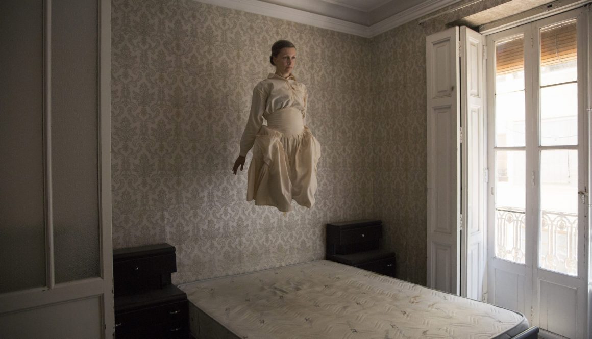 Woman is photographed in a period dress, in an old abandoned house. She appears to be levitating above a bed
