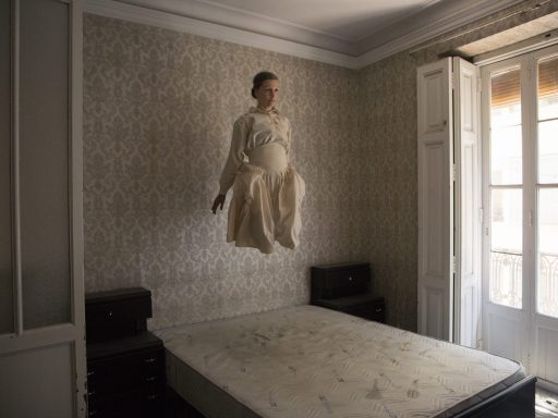 Woman is photographed in a period dress, in an old abandoned house. She appears to be levitating above a bed